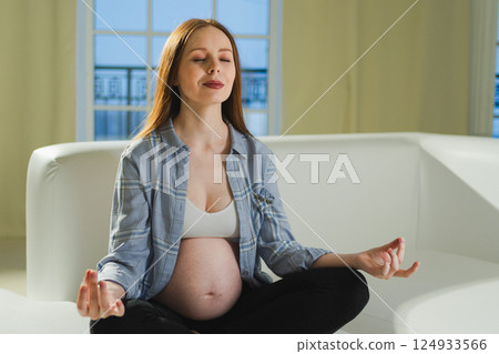 Pregnancy prenatal breathing. Pregnant woman with big belly sitting in lotus pose on sofa at home. Pregnant girl practicing yoga mindfulness meditation. Relaxation self care during pregnancy 124933566