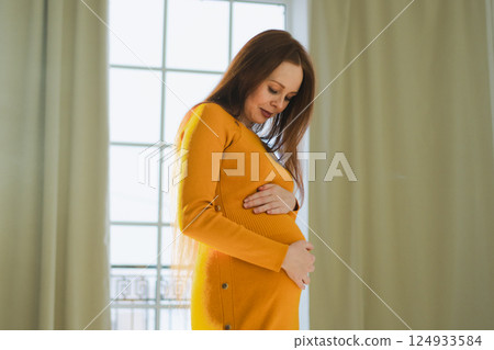 Pregnancy motherhood people expectation future. Pregnant woman with big belly standing near window at home. Girl hugging her tummy enjoying pregnancy. Maternity tenderness parenthood new life concept 124933584