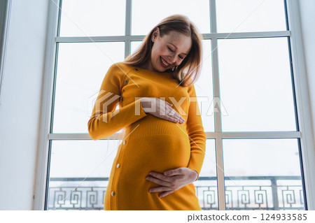 Pregnancy motherhood people expectation future. Pregnant woman with big belly standing near window at home. Girl hugging her tummy enjoying pregnancy. Maternity tenderness parenthood new life concept 124933585