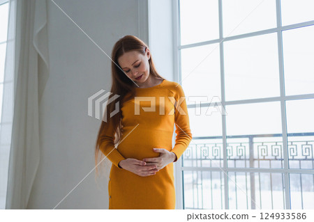Pregnancy motherhood people expectation future. Pregnant woman with big belly standing near window at home. Girl hugging her tummy enjoying pregnancy. Maternity tenderness parenthood new life concept 124933586