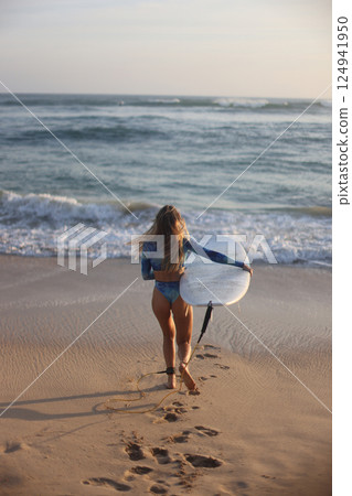 A woman is leisurely walking along the beach while holding a surfboard 124941950