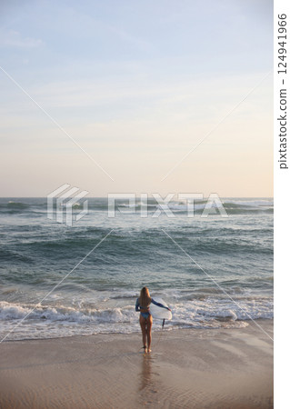 A woman is leisurely walking along the beach while holding a surfboard A woman is leisurely walking along the beach while holding a surfboard 124941966