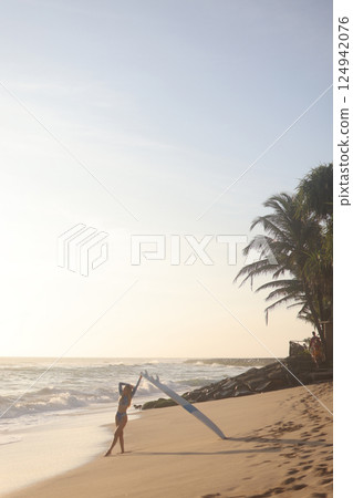 A person is standing on a sandy beach while holding a surfboard 124942076
