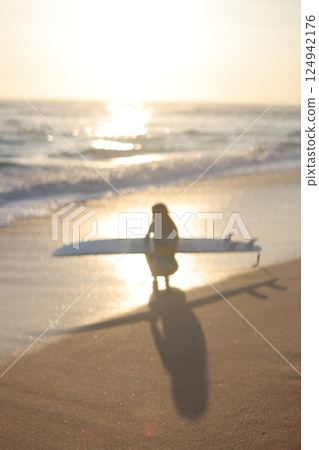 A Surfer Woman Can Be Seen Strolling Along the Beach as the Sun Sets in the Beautiful Sky 124942176