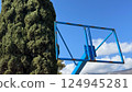 Rusty, hollow blue basketball hoop frame against tall evergreen tree and sky 124945281