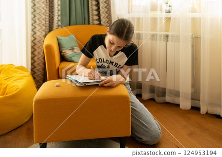Young girl writing in a notebook while sitting on the floor in a cozy living room with colorful 124953194