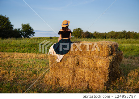 Young beautiful woman in a straw hat looks at the setting sun, the girl sits on a large pile of straw bales. Young beautiful woman in a straw hat looks at the setting sun, the girl sits on a large pile of straw bales. 124953787