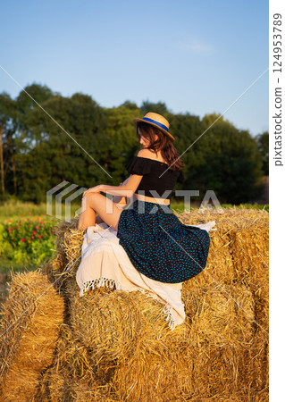 Young beautiful woman in a straw hat looks at the setting sun, the girl sits on a large pile of straw bales. Warm summer evening. Young beautiful woman in a straw hat looks at the setting sun, the girl sits on a large pile of straw bales. Warm summer evening. 124953789
