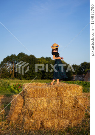 A young beautiful woman in a straw hat stands on a large pile of straw bales. Freedom, wind, setting sun. A young beautiful woman in a straw hat stands on a large pile of straw bales. Freedom, wind, setting sun. 124953790