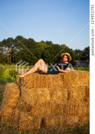 A young beautiful woman in a straw hat lies on a large pile of straw bales and looks at the setting sun. A young beautiful woman in a straw hat lies on a large pile of straw bales and looks at the setting sun. 124953791