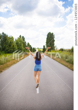 Attractive girl, stands posing, hands up in the middle of an asphalt road near the field, copies space for content. Attractive girl, stands posing, hands up in the middle of an asphalt road near the field, copies space for content. 124953803