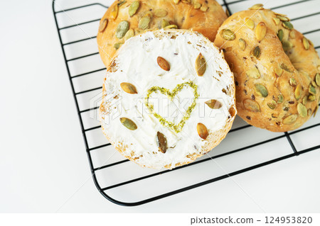 Delicious pumpkin seed bagels topped with cream cheese and a heart-shaped sprinkle design, displayed on a wire rack 124953820