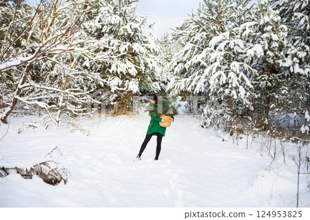 A young beautiful girl in a green sweater and hat stands in the winter forest along with a basket with pine branches. 124953825
