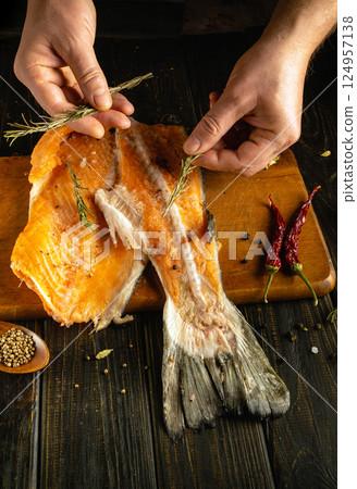 Fresh salmon fillet rests on a wooden cutting board as hands sprinkle herbs and spices. Red chilies and peppercorns enhance the rustic kitchen atmosphere, emphasizing culinary preparation 124957138