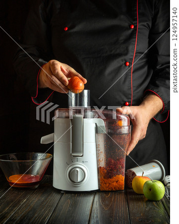 A person wearing a black apron is juicing fresh apples in a cozy kitchen. The juicer is prominently featured alongside bowls full of apple juice and pulp on a wooden countertop 124957140