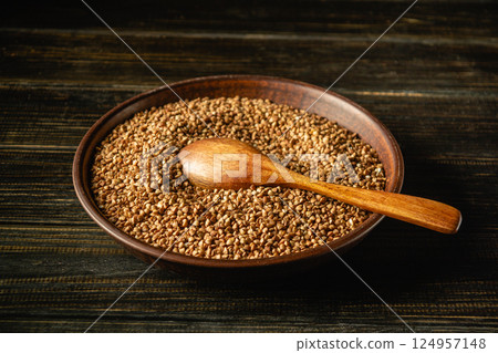 A close-up of a wooden bowl filled with brown buckwheat seeds. A wooden spoon rests on top of the buckwheat grains, creating a rustic and natural atmosphere, perfect for culinary use A close-up of a wooden bowl filled with brown buckwheat seeds. A wooden spoon rests on top of the buckwheat grains, creating a rustic and natural atmosphere, perfect for culinary use 124957148
