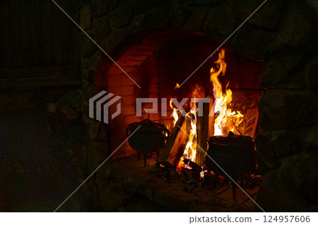 Open fire in the fireplace. A stove in which food is cooked. Cast-iron cauldrons for cooking at the stake. Firewood in the fireplace. Cozy home atmosphere. Night. Open fire in the fireplace. A stove in which food is cooked. Cast-iron cauldrons for cooking at the stake. Firewood in the fireplace. Cozy home atmosphere. Night. 124957606