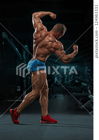 muscular man flexes his biceps and showcases his well defined physique while standing in gym setting. low light highlights his strong body as he exercises. 124963331
