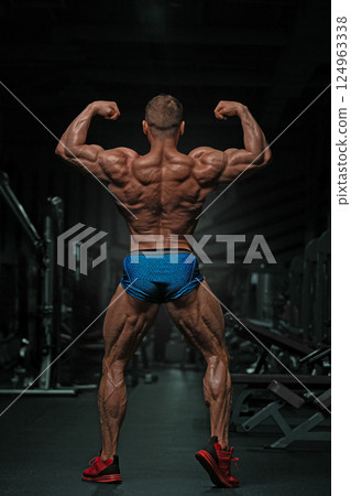 athletic man demonstrates his muscular build while flexing in gym. setting is dimly lit highlighting his physique against workout equipment and creating intense atmosphere. 124963338