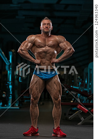 athletic man stands proudly in gym flexing his muscles in striking pose. environment is equipped various fitness machines emphasizing strength and dedication. 124963340