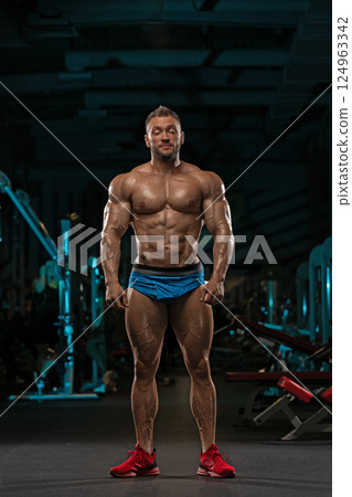 athletic man stands proudly in gym highlighting his well defined muscles as he prepares for workout. dimly lit environment emphasizes his impressive physique. athletic man stands proudly in gym highlighting his well defined muscles as he prepares for workout. dimly lit environment emphasizes his impressive physique. 124963342
