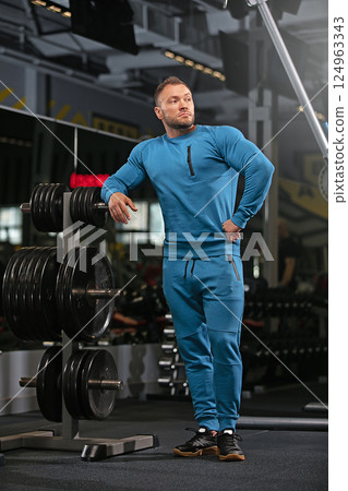 athletic man stands in gym wearing blue workout attire. He leans against weight rack in fitness center showcasing strength and confidence. Bright lighting enhances setting. athletic man stands in gym wearing blue workout attire. He leans against weight rack in fitness center showcasing strength and confidence. Bright lighting enhances setting. 124963343