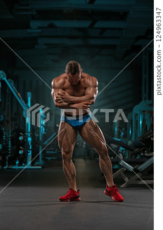 Muscular man stands in gym flexing his arms while wearing blue shorts and red shoes. atmosphere is intense dim lighting emphasizing his well defined muscles. 124963347
