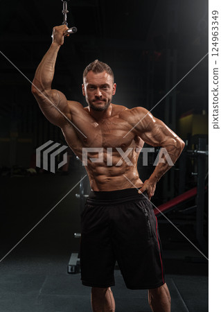 Well built man shows off his muscular physique in gym environment posing cable machine. lighting accentuates his toned muscles during strength training session. 124963349