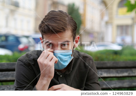 Young man sits on the bench in park and puts on medical protective face mask 124963829