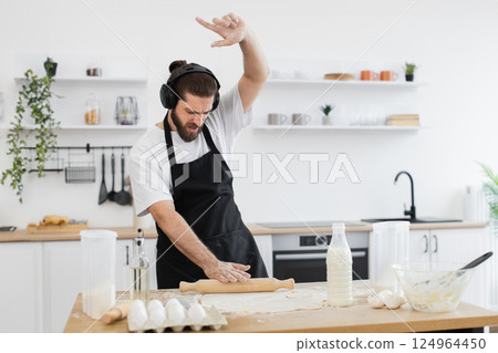Bearded young adult male wearing black apron and headphones baking in bright kitchen, rolling dough enthusiastically while listening to music. 124964450
