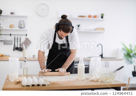 Man wearing headphones and apron rolls dough with rolling pin on kitchen table. Modern kitchen setting with baking ingredients. Man appears to be in twenties demonstrating multitasking activity. Man wearing headphones and apron rolls dough with rolling pin on kitchen table. Modern kitchen setting with baking ingredients. Man appears to be in twenties demonstrating multitasking activity. 124964451