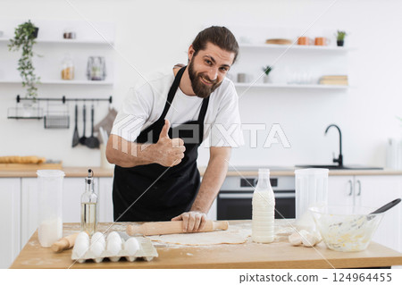Smiling young adult male chef with dark hair rolling dough in bright kitchen while giving thumbs up gesture. Ingredients including eggs, milk, and flour are neatly arranged in foreground on table. 124964455