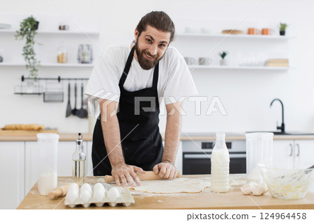 Young adult man wearing black apron in modern kitchen rolling out dough, surrounded by baking ingredients, appearing happy and engaged in food preparation. 124964458