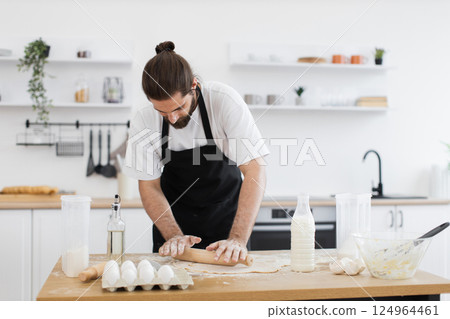 Adult male baker making dough in bright home kitchen environment with cooking tools and ingredients on wooden table. Creative baking process featuring young man wearing apron. 124964461