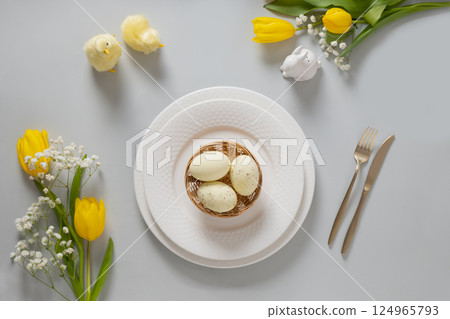 Easter table setting with spring yellow tulips, white plates, bunny, eggs on gray background. Happy Easter celebration. Copy space. View from above. Easter table setting with spring yellow tulips, white plates, bunny, eggs on gray background. Happy Easter celebration. Copy space. View from above. 124965793