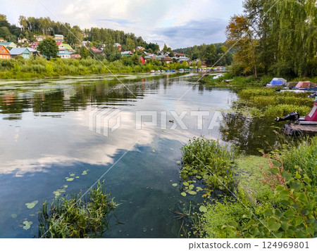Village lake with houses and boats on the shore, banks overgrown with reeds, summer rural calm landscape Village lake with houses and boats on the shore, banks overgrown with reeds, summer rural calm landscape 124969801