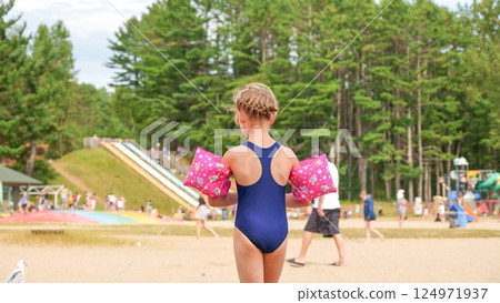Little girl with pink water wings standing on the sandy beach near the playground 124971937