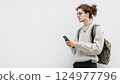 Young person in a gray sweater with backpack using smartphone against a plain white background 124977796