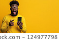 Excited young man holds smartphone with yellow background, showcasing joy and surprise 124977798