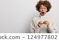 Surprised woman with curly hair checking smartphone and expressing excitement in casua 124977802