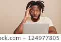 Concerned male with dreadlocks experiences surprise and disbelief while working on a laptop at home 124977805