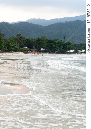 Soft waves kiss the sandy shore while tropical greenery and distant mountains create a serene oasis. 124978340