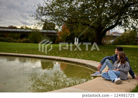 Romantic young couple relaxing by the pond in autumn park, wearing cozy casual outfits, enjoying a peaceful moment together. Romantic young couple relaxing by the pond in autumn park, wearing cozy casual outfits, enjoying a peaceful moment together. 124981725