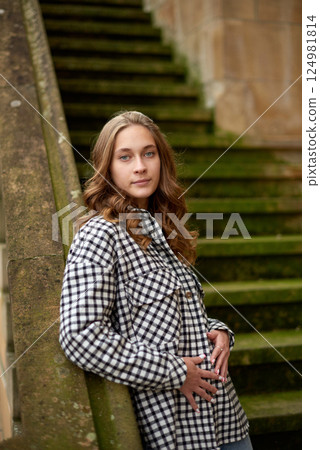 Young woman in casual checkered outfit posing on mossy stone staircase in a charming European village with red shutter windows and rustic architecture. Vibrant European Garden Scene: Carefree Young 124981814