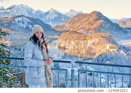 Beautiful Young Woman Standing in Snowy Winter Landscape with Mountain and Lake Backdrop Under Golden Light 124981921