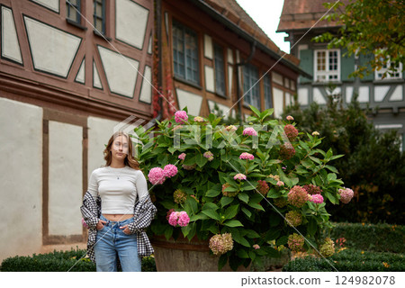 Young woman posing with hydrangeas in a European garden, surrounded by half-timbered houses and autumn foliage, expressing joy and elegance. 124982078