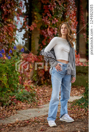 Stylish young woman in trendy autumn outfit posing confidently in a park with vibrant fall foliage, wearing a black-and-white plaid shirt over a white crop top, paired with blue jeans and white 124982085