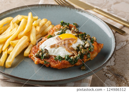 Delicious breaded schnitzel with anchovies, fried egg and capers parsley sauce on top served with french fries close-up on a plate. Horizontal 124986312