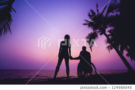 Disabled man in a wheelchair with his wife on the beach. Silhouettes at sunset Disabled man in a wheelchair with his wife on the beach. Silhouettes at sunset 124986848