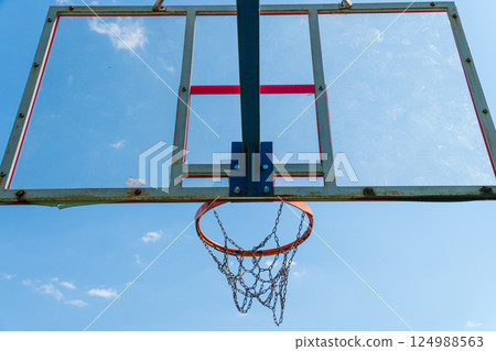 A weathered basketball hoop and net are positioned against a bright blue sky, showcasing a peaceful afternoon atmosphere 124988563
